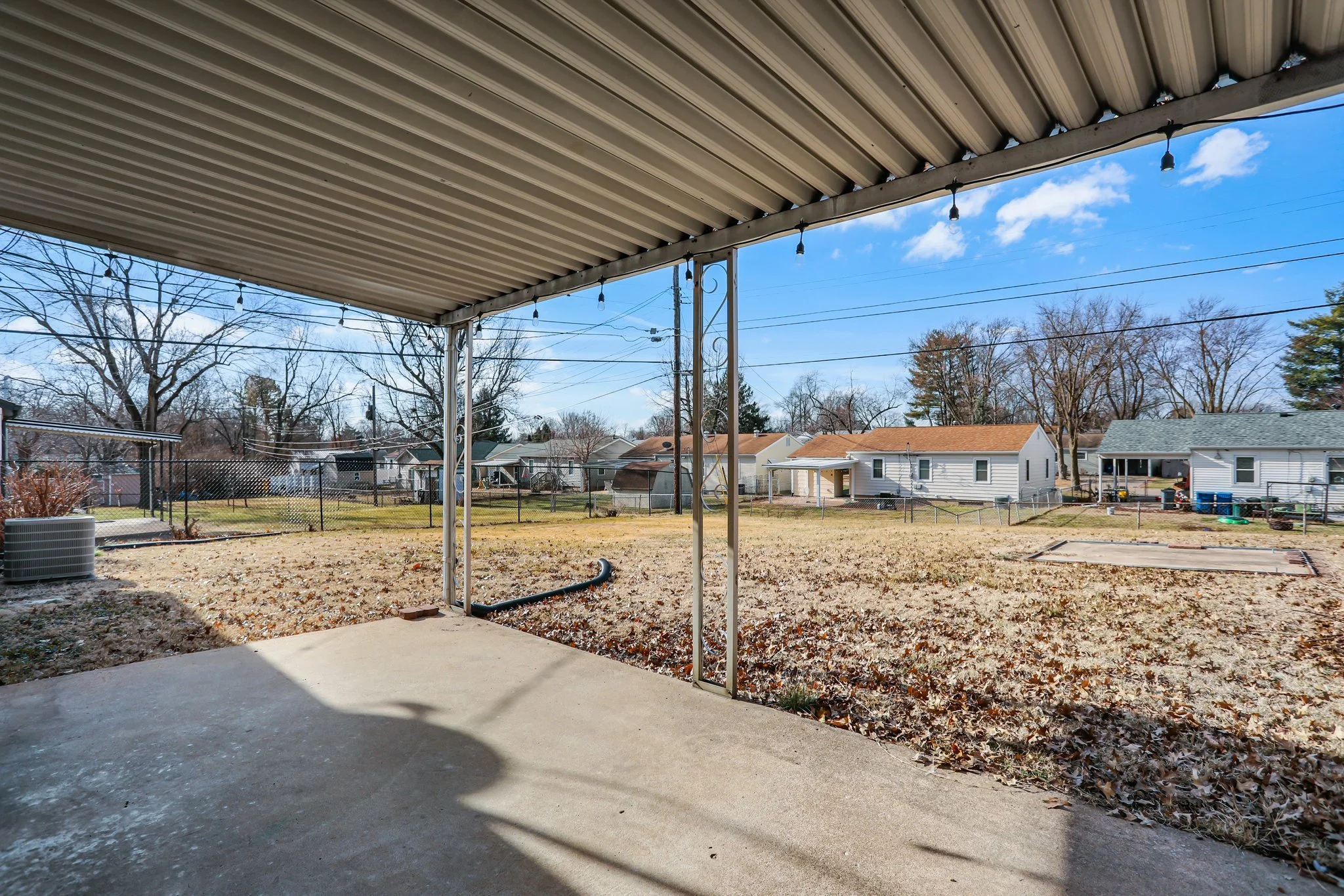 Backyard with a covered patio and string lights, in a suburban neighborhood with houses, trees, and power lines under a blue sky.