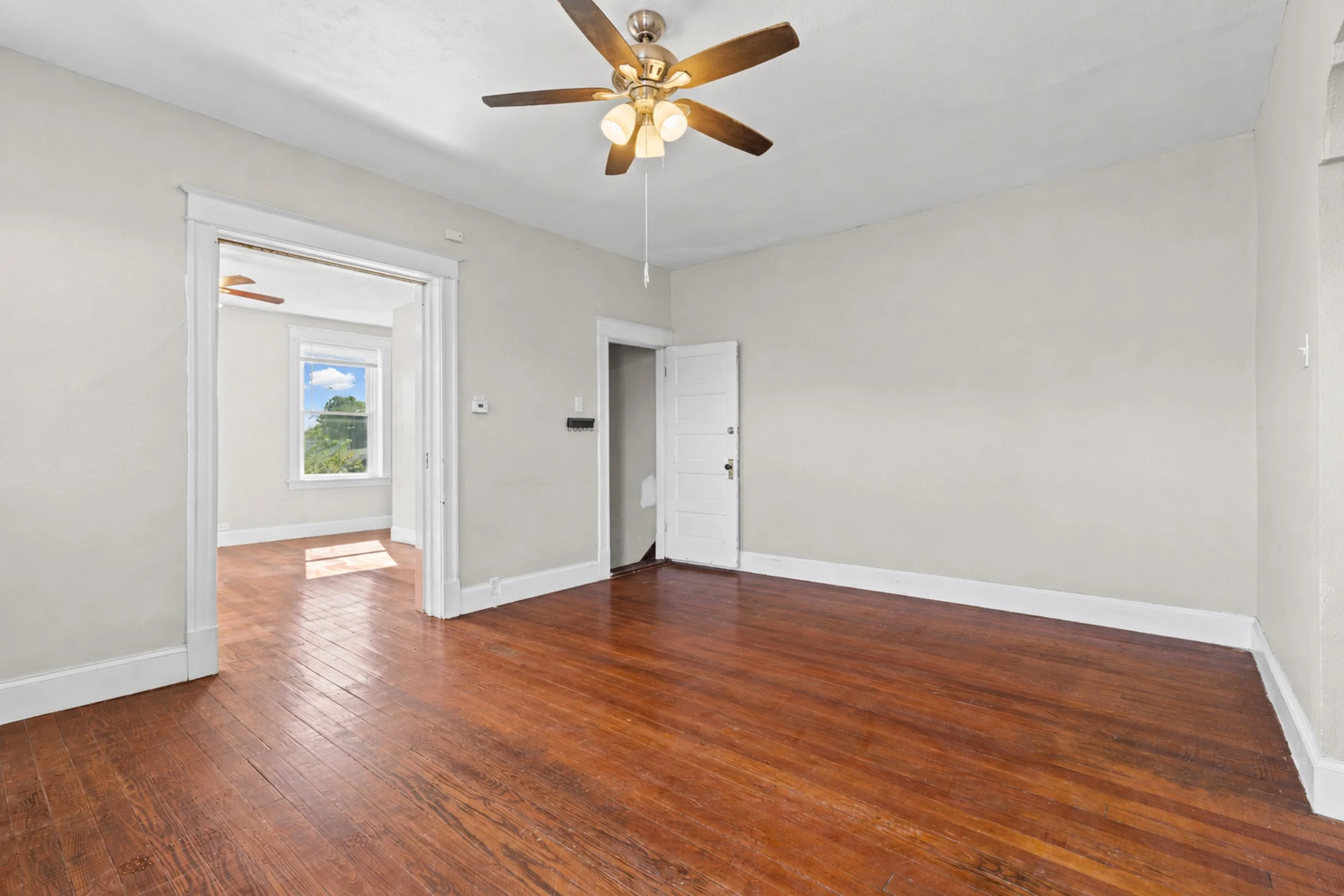 Empty room with hardwood floors, white walls, ceiling fan with lights, open door, window showing greenery outdoors.