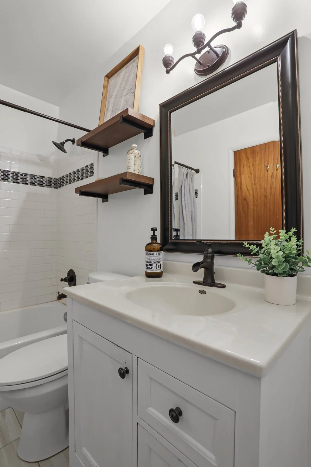 Bathroom with white vanity, black faucet, large mirror, small potted plant, soap dispenser, bathtub, showerhead, and wooden shelves with decor and toiletries.