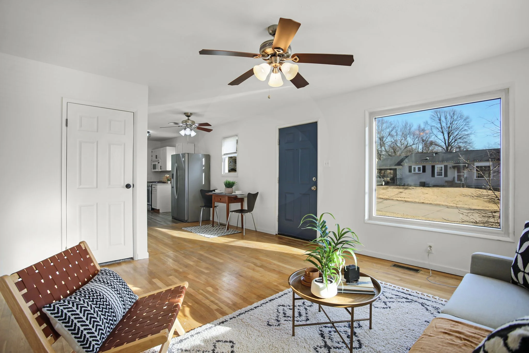 Bright living room with hardwood floors, large windows, ceiling fans, and minimal furniture including a woven lounge chair, a small coffee table with plants, and a sofa. A view of a yard with a house and trees outside.