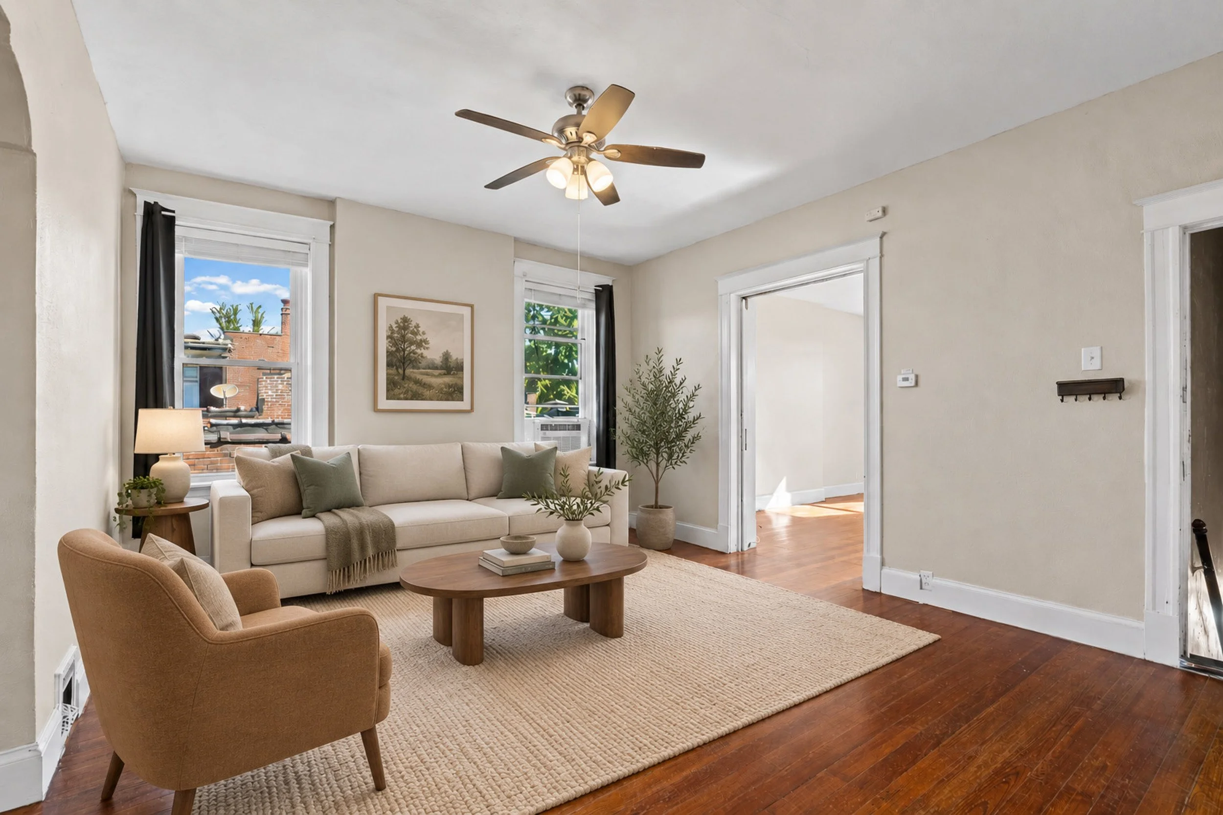 Living room with a beige sofa, a round wooden coffee table, an armchair, and plants near large windows with black curtains, hardwood floors, and a ceiling fan.