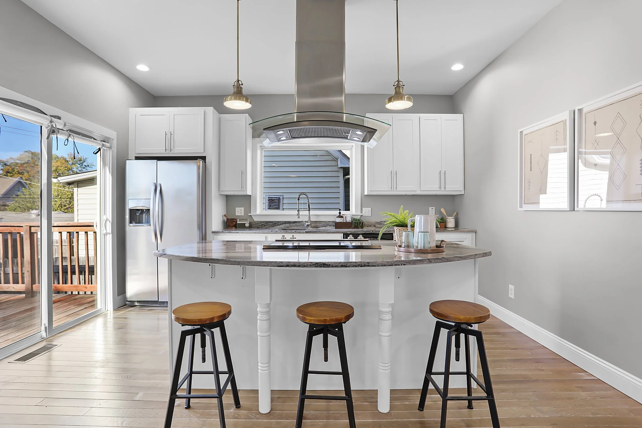 A modern kitchen with white cabinets, a stainless steel refrigerator, a kitchen island with a granite countertop, three wooden barstools, a sliding glass door leading to a deck, and a window above the sink letting in natural light. There are two pend