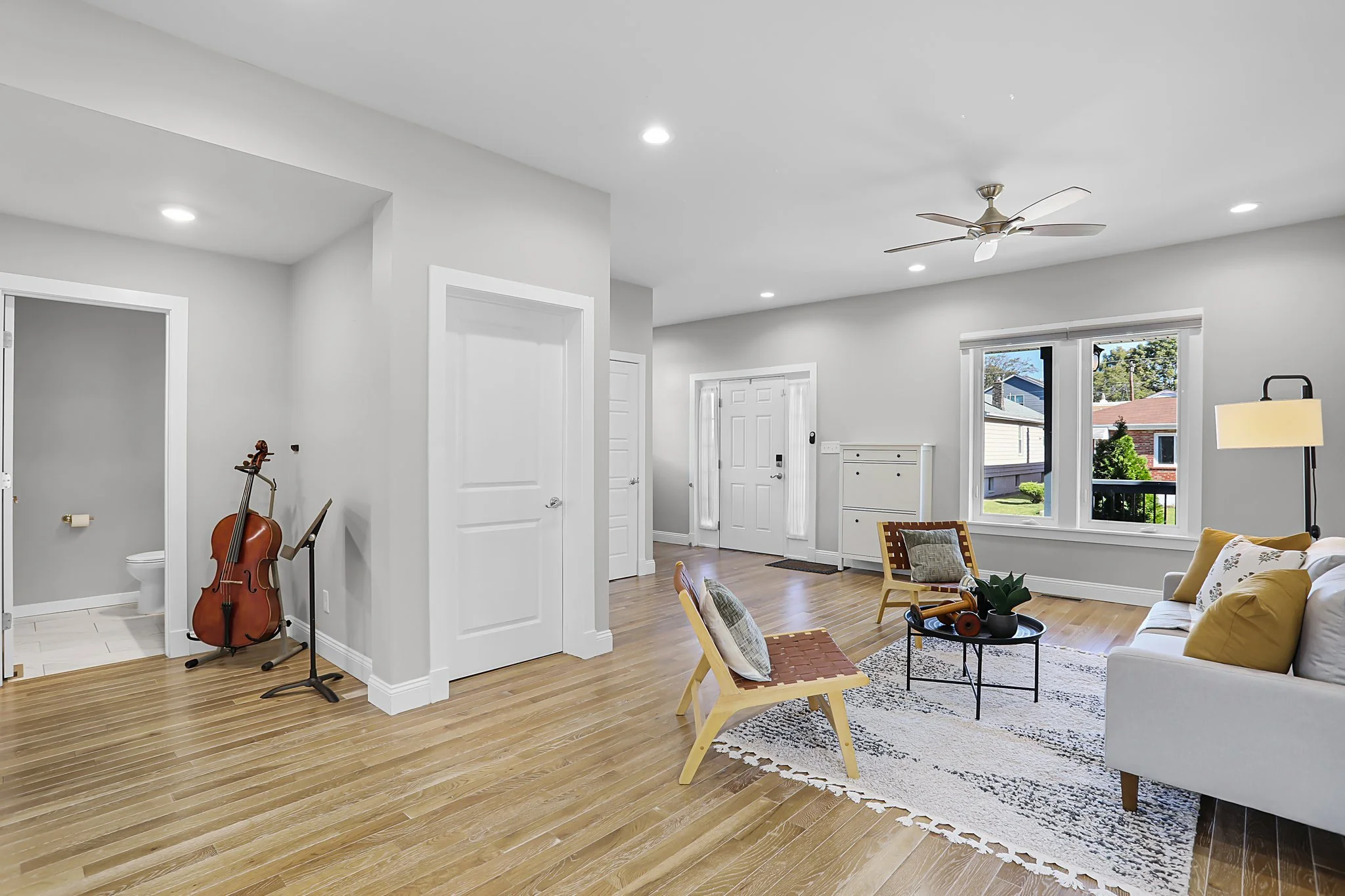 A bright living room with white walls, hardwood floors, and large windows showing a blue sky and greenery outside. There is a white sofa with yellow and floral cushions, a wooden chair with a woven seat, and a small round black table with a plant and