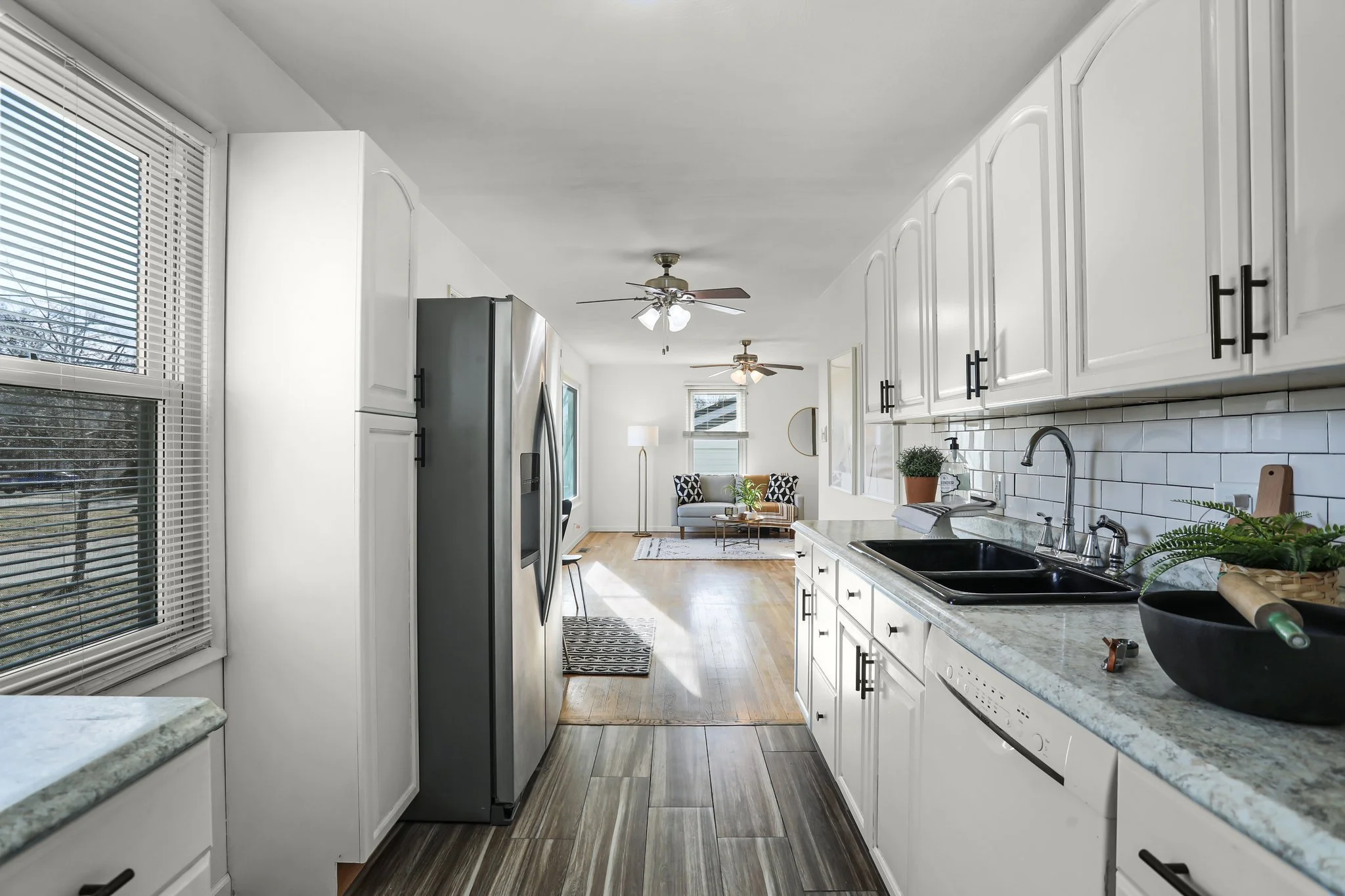 Bright white kitchen with white cabinets, black hardware, stainless steel refrigerator, granite countertops, sink with plants, and view into living room with ceiling fans, large windows, and a small sofa.