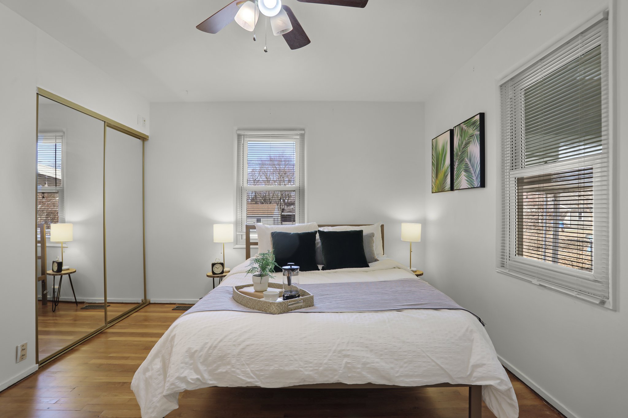 A bedroom with white walls and large windows. There is a bed with white bedding and black pillows. Two bedside tables with lamps are on either side of the bed. A tray with a plant and a French press is on the bed. A closet with sliding mirror doors a