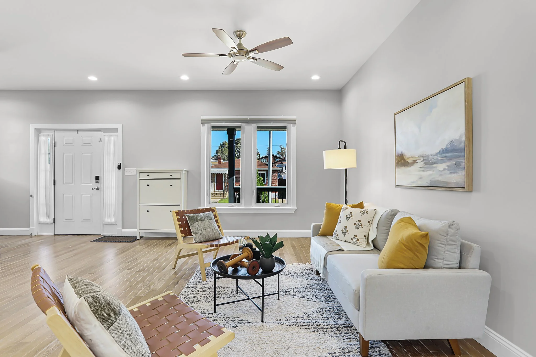 Bright living room with white walls, hardwood floors, a white sofa with yellow pillows, a large abstract painting, a floor lamp, woven chairs, a small table with decorative items, a window showing a neighborhood view, and a ceiling fan.
