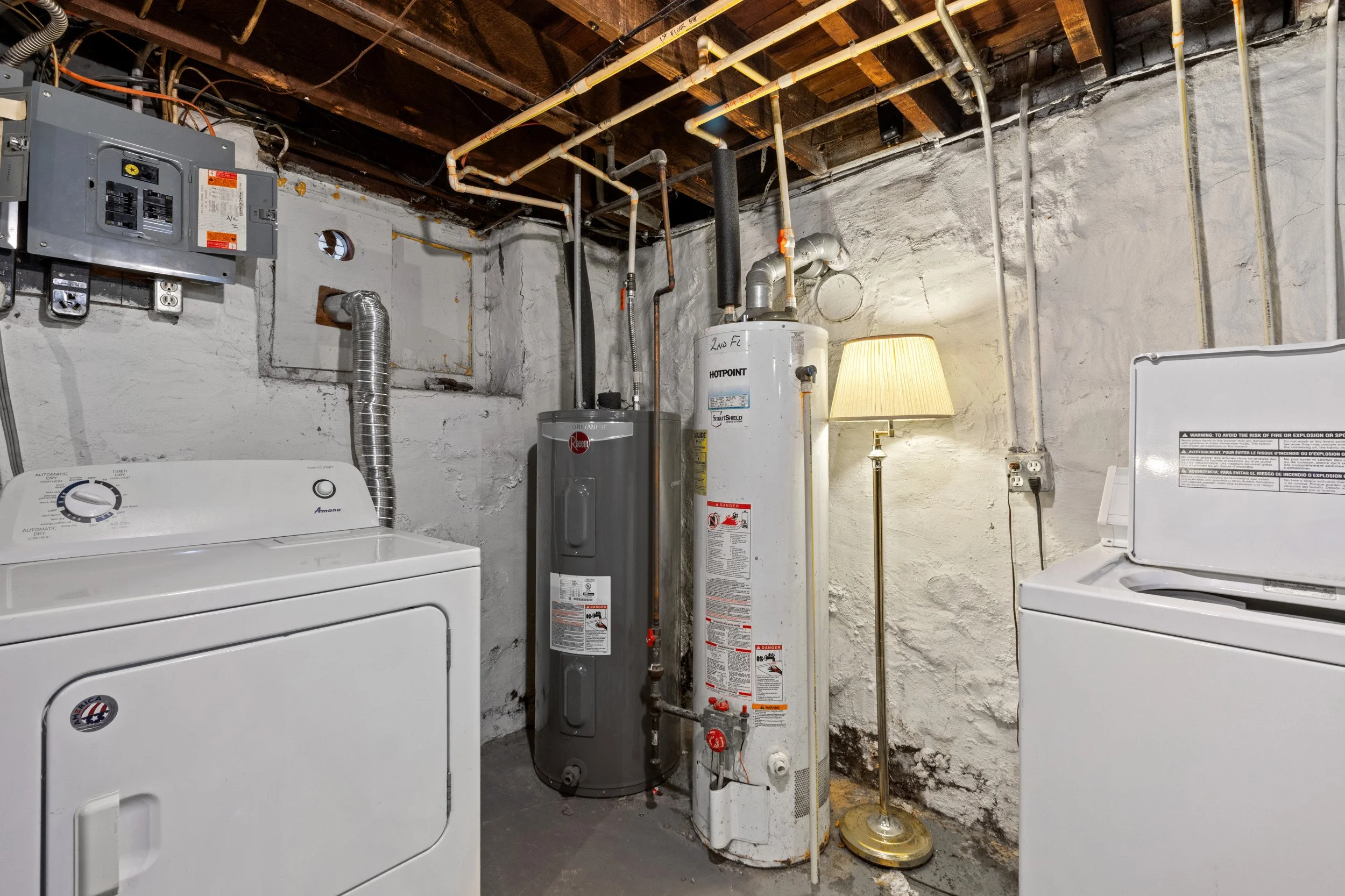 A laundry room with a washing machine, a water heater, a lamp, and various pipes and electrical panels mounted on an unfinished white brick wall with exposed wooden beams on the ceiling.