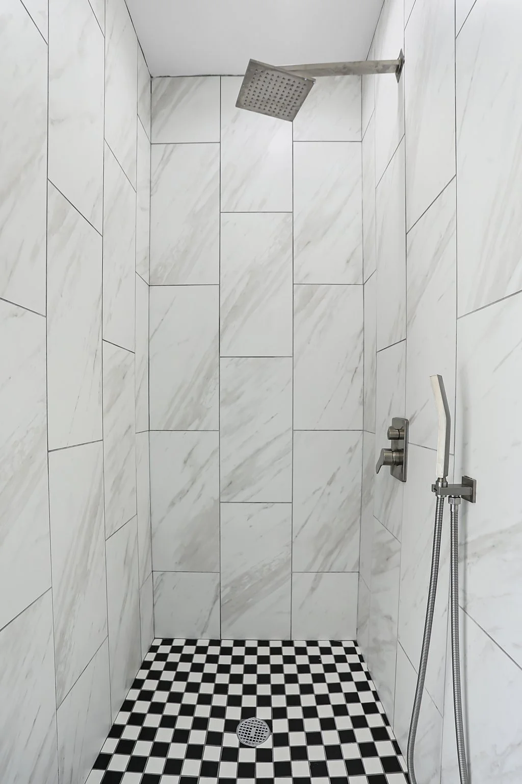 A modern walk-in shower with white marble tiles on the walls, black-and-white checkered tile flooring, a square overhead showerhead, a handheld showerhead, and a built-in wall control.