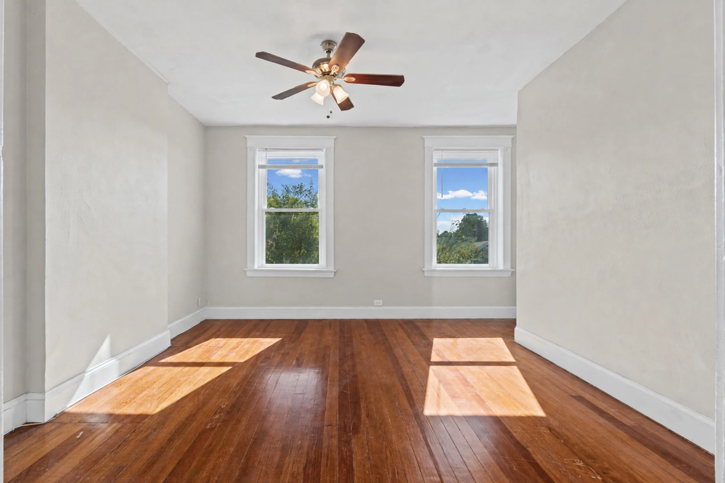Empty room with two windows, white walls, wooden floor, and ceiling fan with lights.