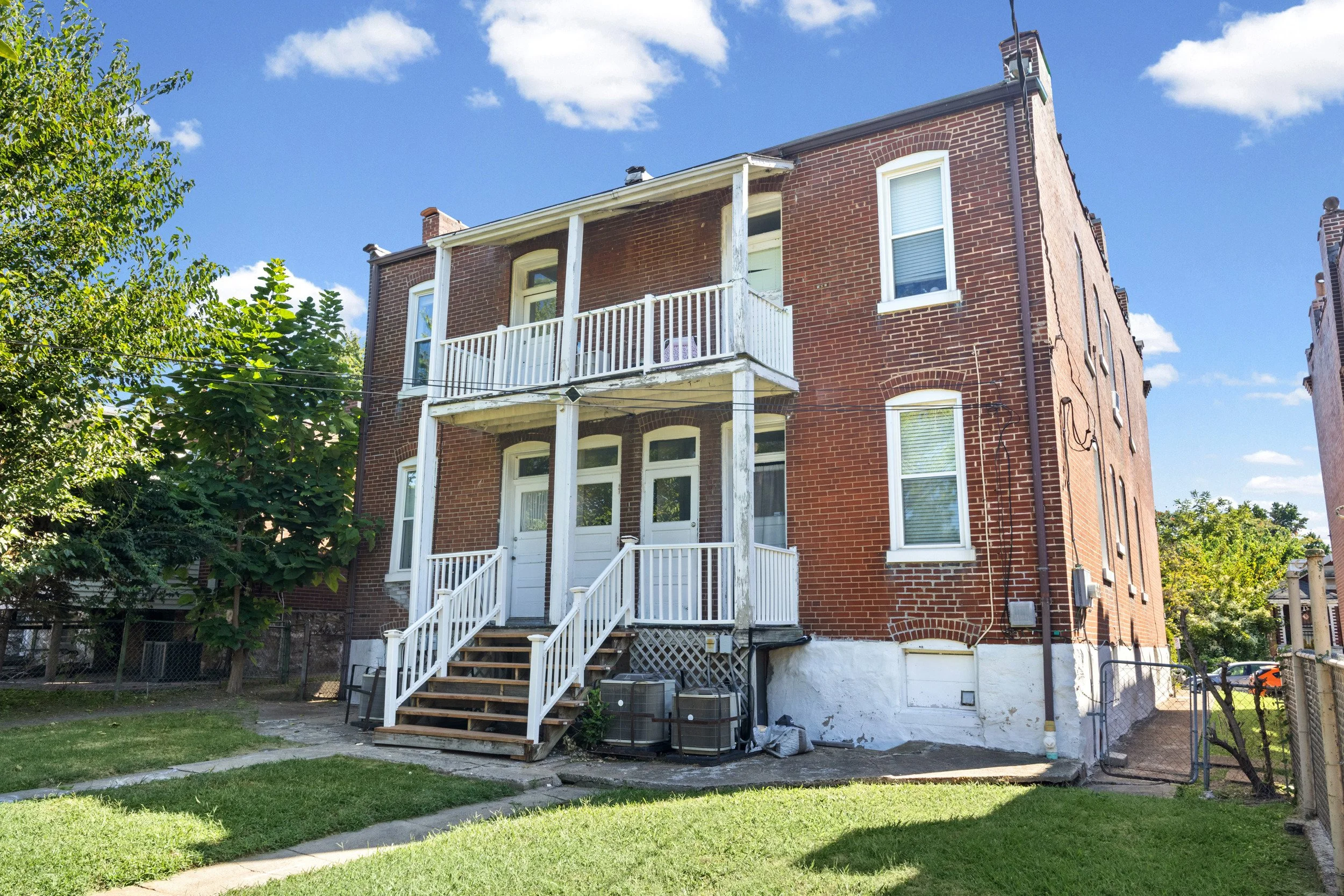 Back of a three-story brick apartment building with a small porch and stairs, surrounded by trees and a grassy yard under a blue sky with clouds.