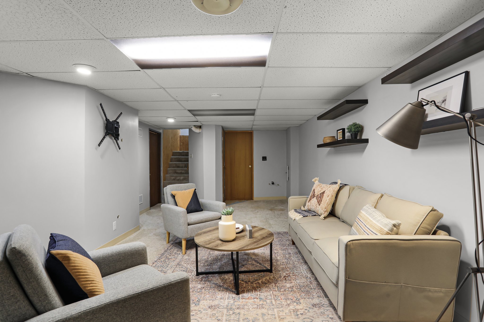 A cozy living room featuring a beige sofa with throw pillows, a round wooden coffee table, two armchairs with pillows, a rug, a wall-mounted TV, and wall shelves with decorative items. The ceiling has a drop-ceiling with fluorescent lights, and a sta