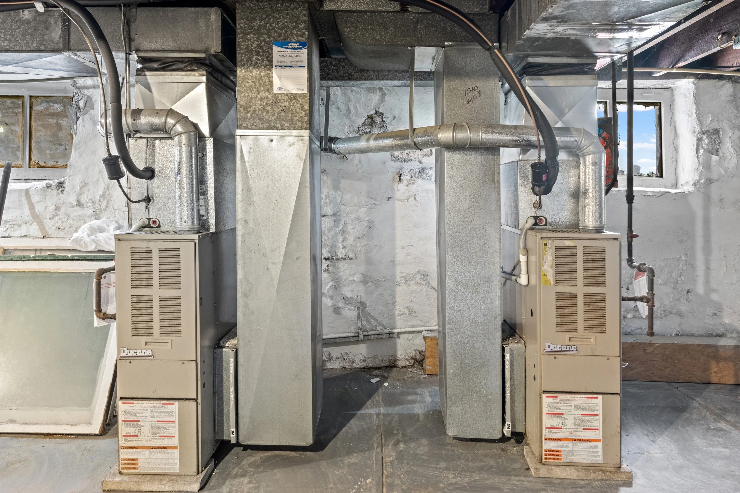 Two dual HVAC units with ductwork and electrical wiring in a partially finished basement or utility room, with small windows in the background.