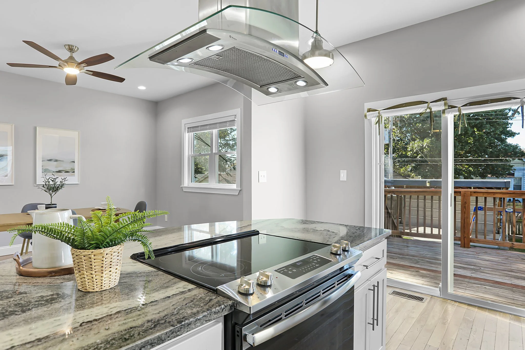 A modern kitchen with a granite countertop stove, a potted plant, and a view of an outdoor deck through sliding glass doors. The kitchen has white cabinets, a stainless steel range hood, and large windows letting in natural light.