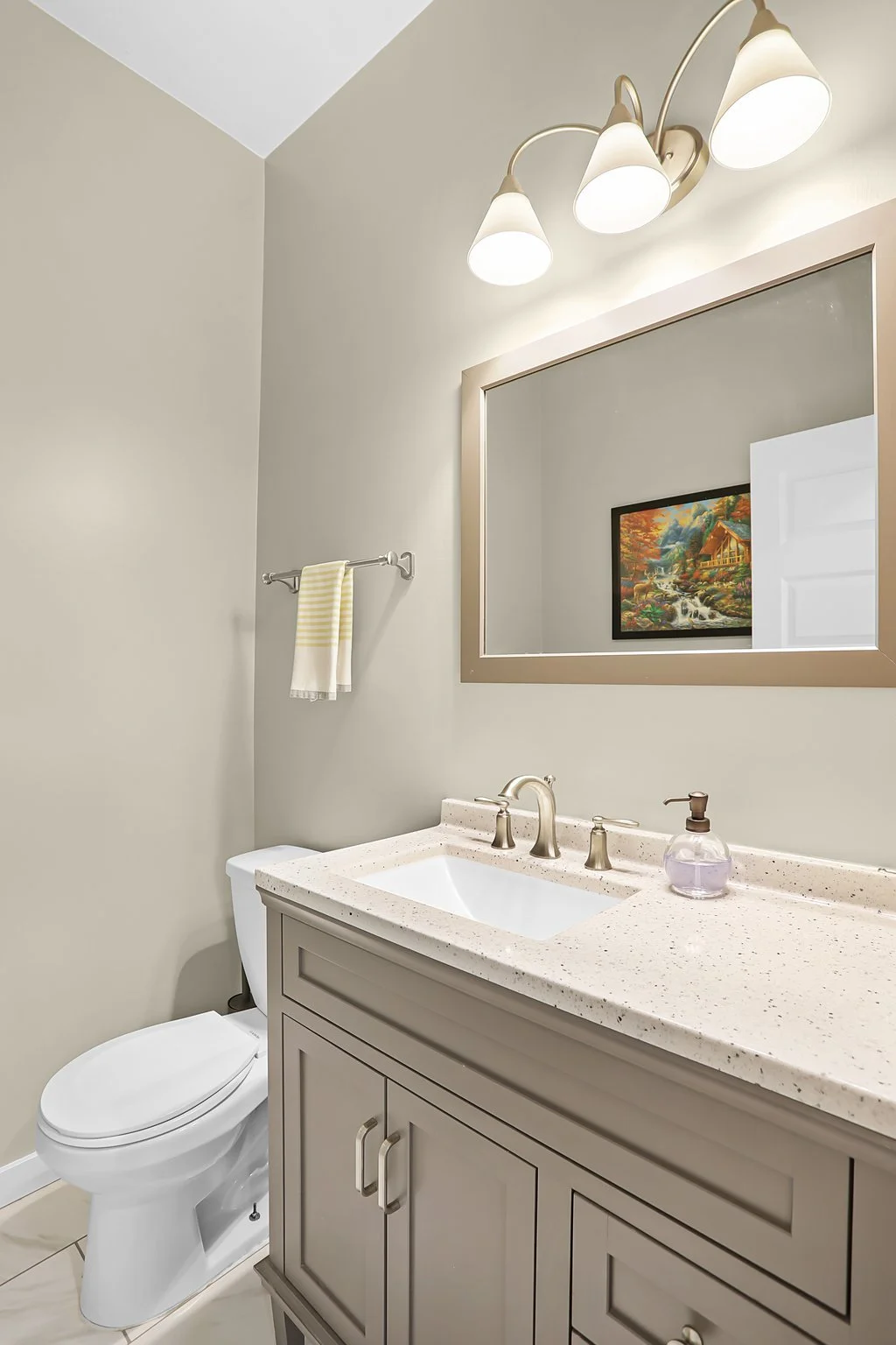 Modern bathroom with beige walls, a large mirror with a metallic frame, a sink with a beige speckled countertop, a silver faucet, a soap dispenser, a towel bar with a yellow and white towel, a framed picture on the opposite wall, and a three-light fi
