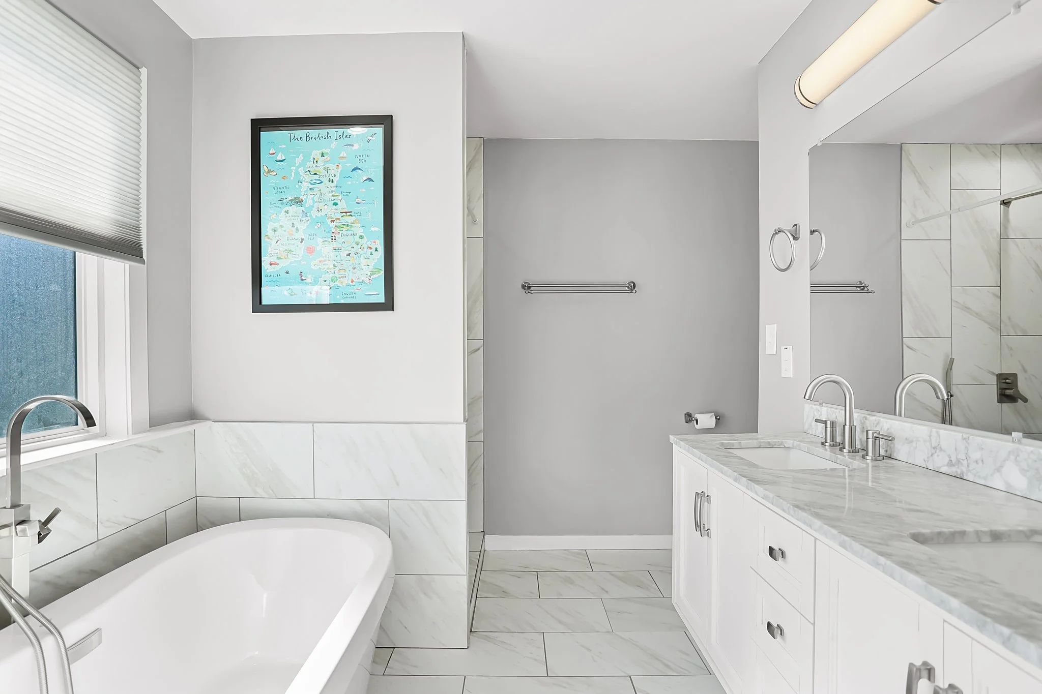 Modern bathroom with white marble countertops and a large mirror above a double sink. A framed illustration of the British Isles is on the wall next to a window with beige blinds. Towel bars and fixtures are made of brushed metal.