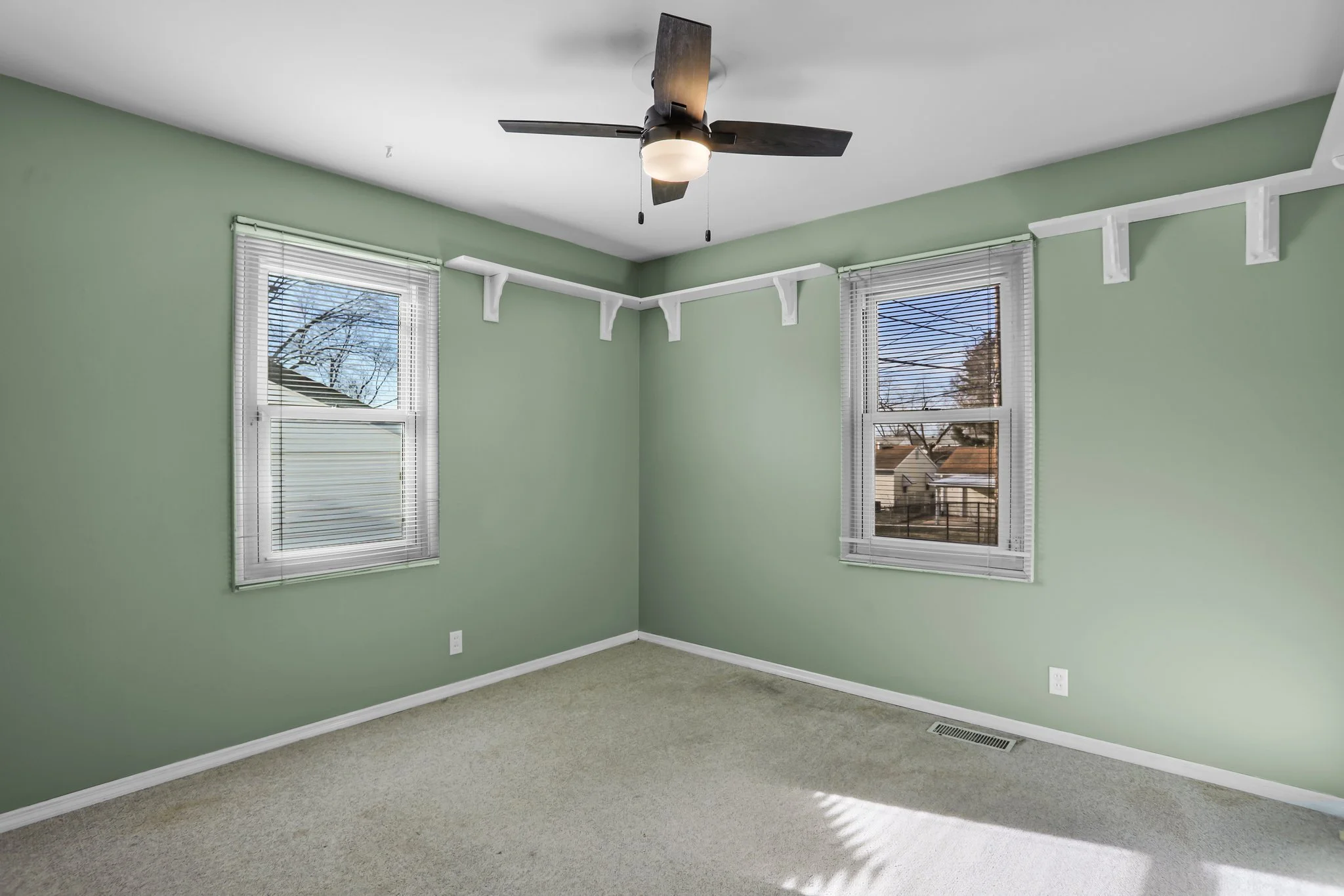 A empty room with green walls, two windows with blinds, a ceiling fan with a light, and beige carpet flooring.