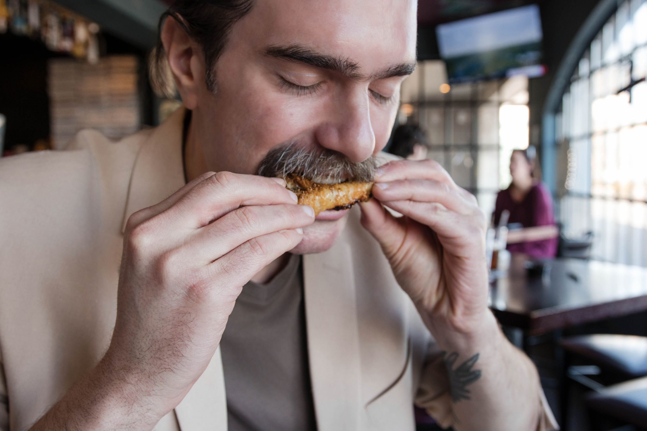 Rueben Wuerdeman with his mustache and a tattoo on his arm, wearing a beige blazer, eating a chicken wing in a restaurant with large windows and other diners in the background.
