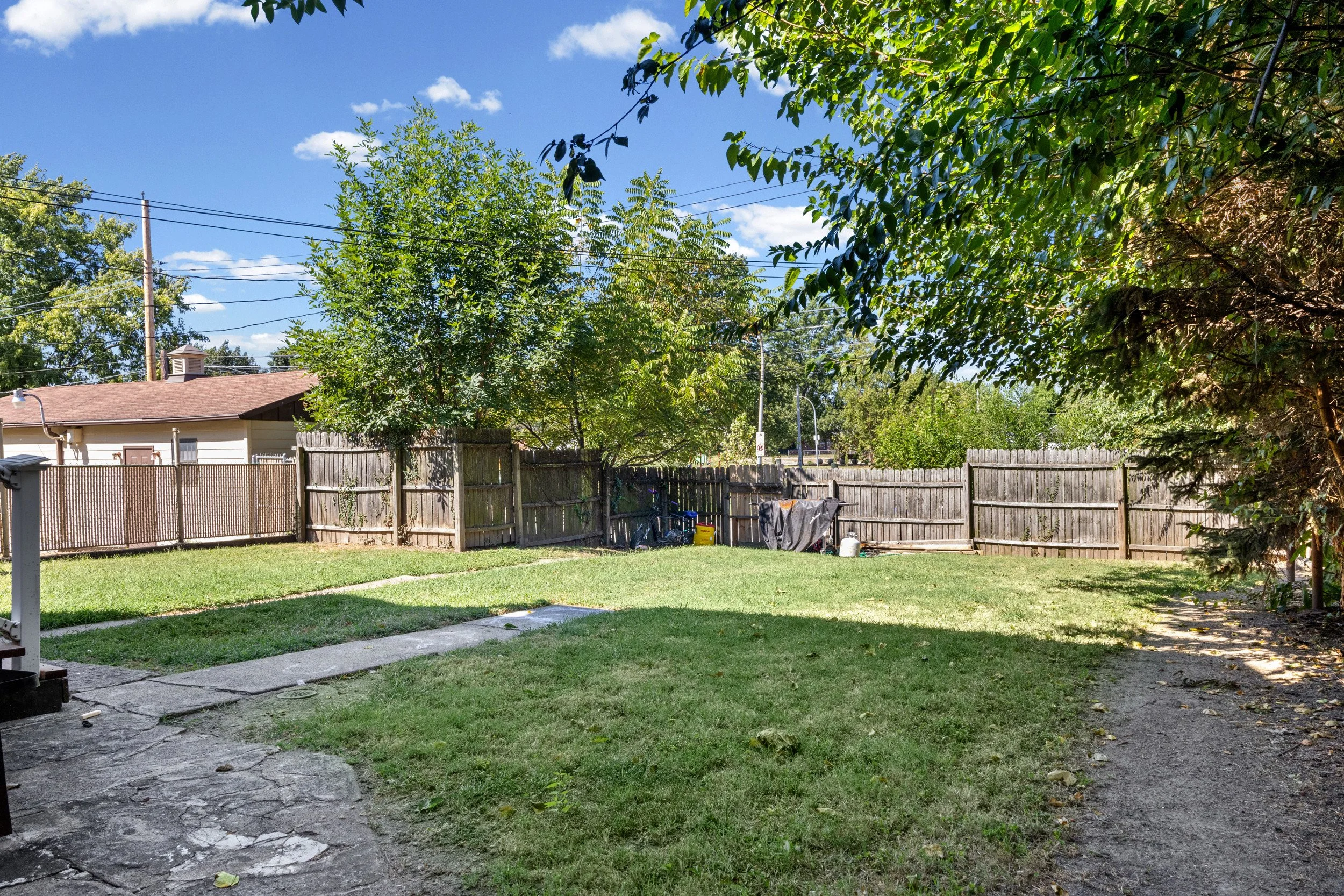 Backyard garden with grass lawn, stone patio, wooden fence, trees, and a house with a brown roof against a blue sky with some clouds.