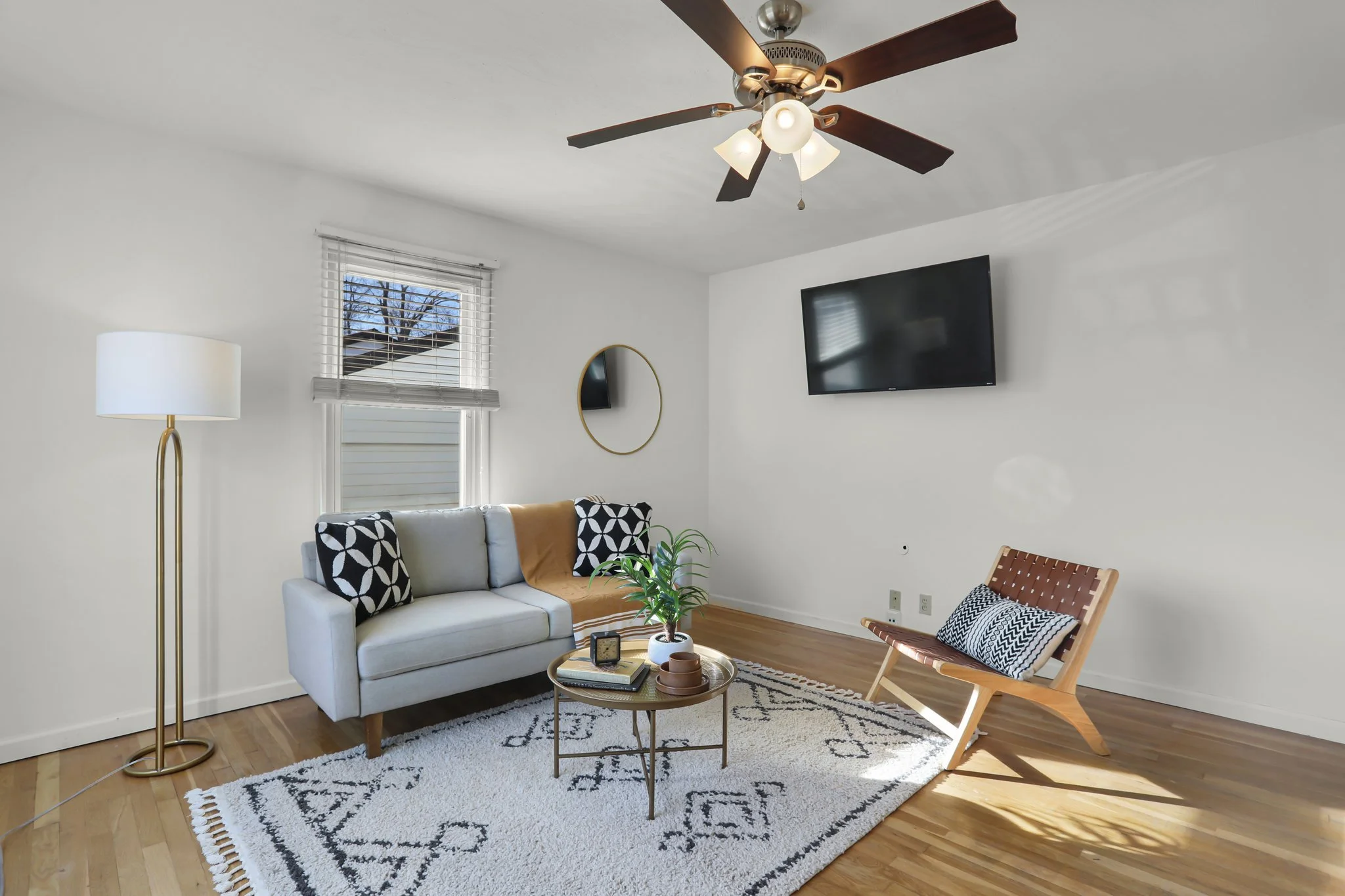 Living room with white walls, a ceiling fan with wooden blades, a wall-mounted flat-screen TV, a round mirror, a tall brass floor lamp with a white lampshade, a beige sofa with patterned pillows, a small round coffee table with books and decor, a pat