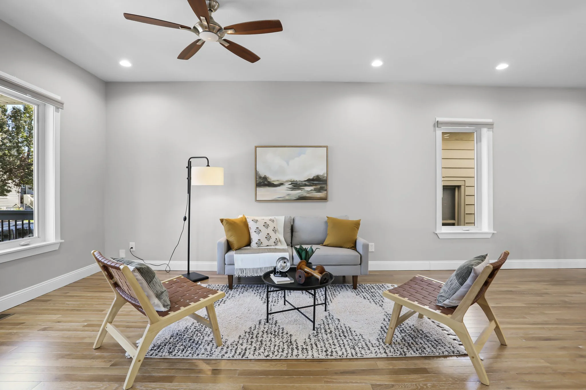 Living room with white sofa, painting on the wall, wooden chairs, a rug, and a side table with decorative items. Two windows and a ceiling fan with wooden blades are visible.