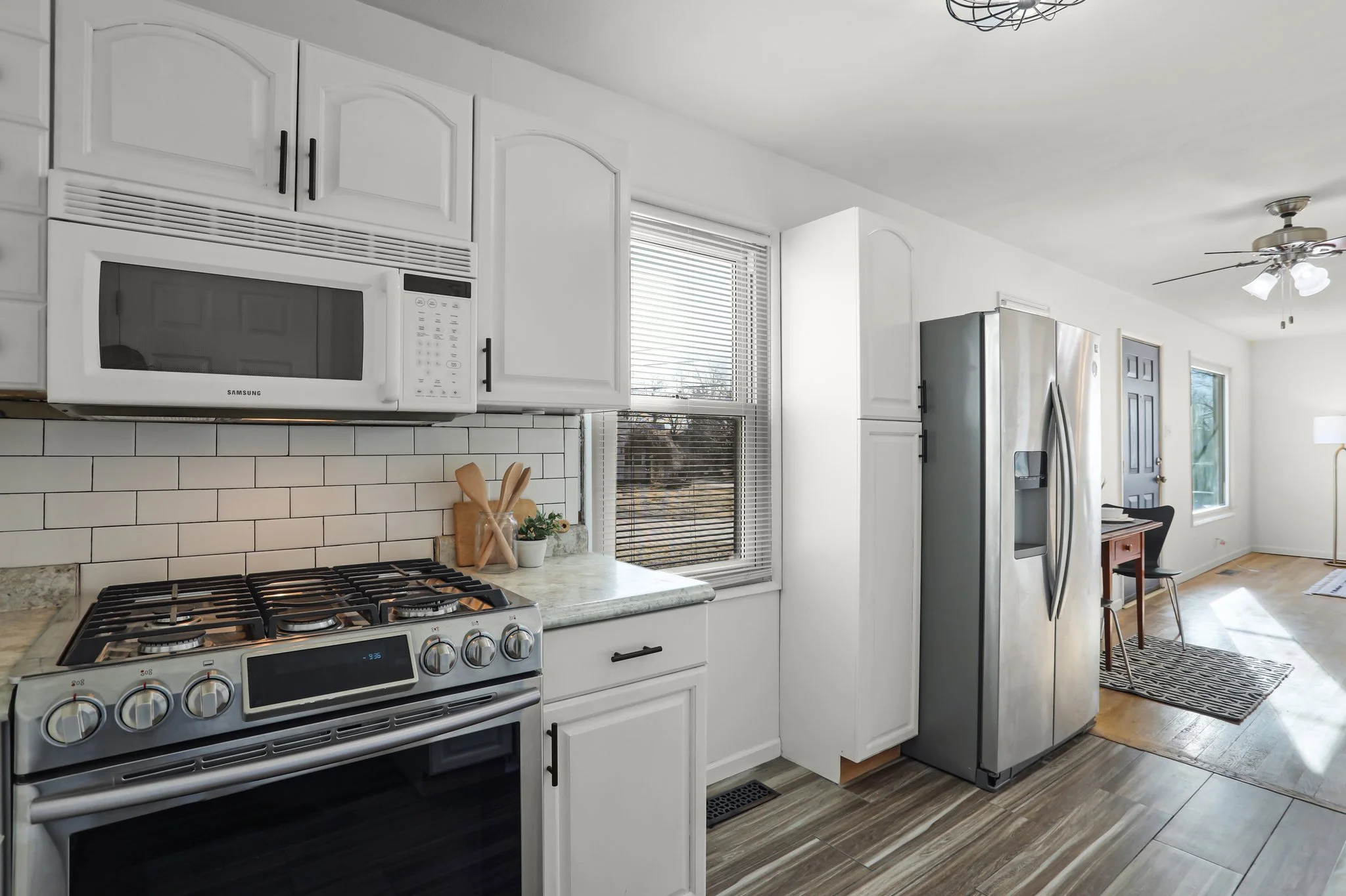 A modern kitchen with white cabinets, a stainless steel microwave, stove, and refrigerator, wood flooring, a window with blinds, and a ceiling fan with lights.