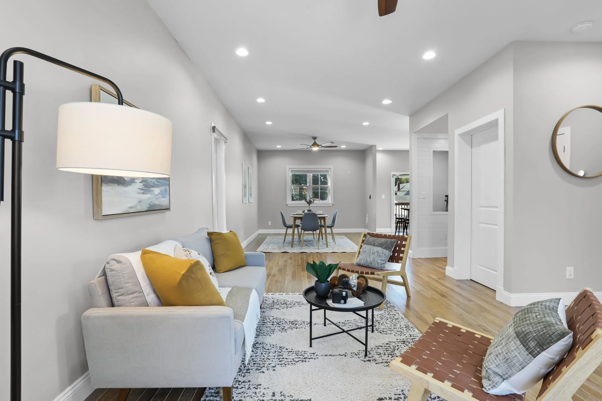 Living room with white couch, yellow pillows, wooden chairs, modern artwork, dining area with a table and four chairs, hardwood flooring, ceiling fan, windows with natural light.