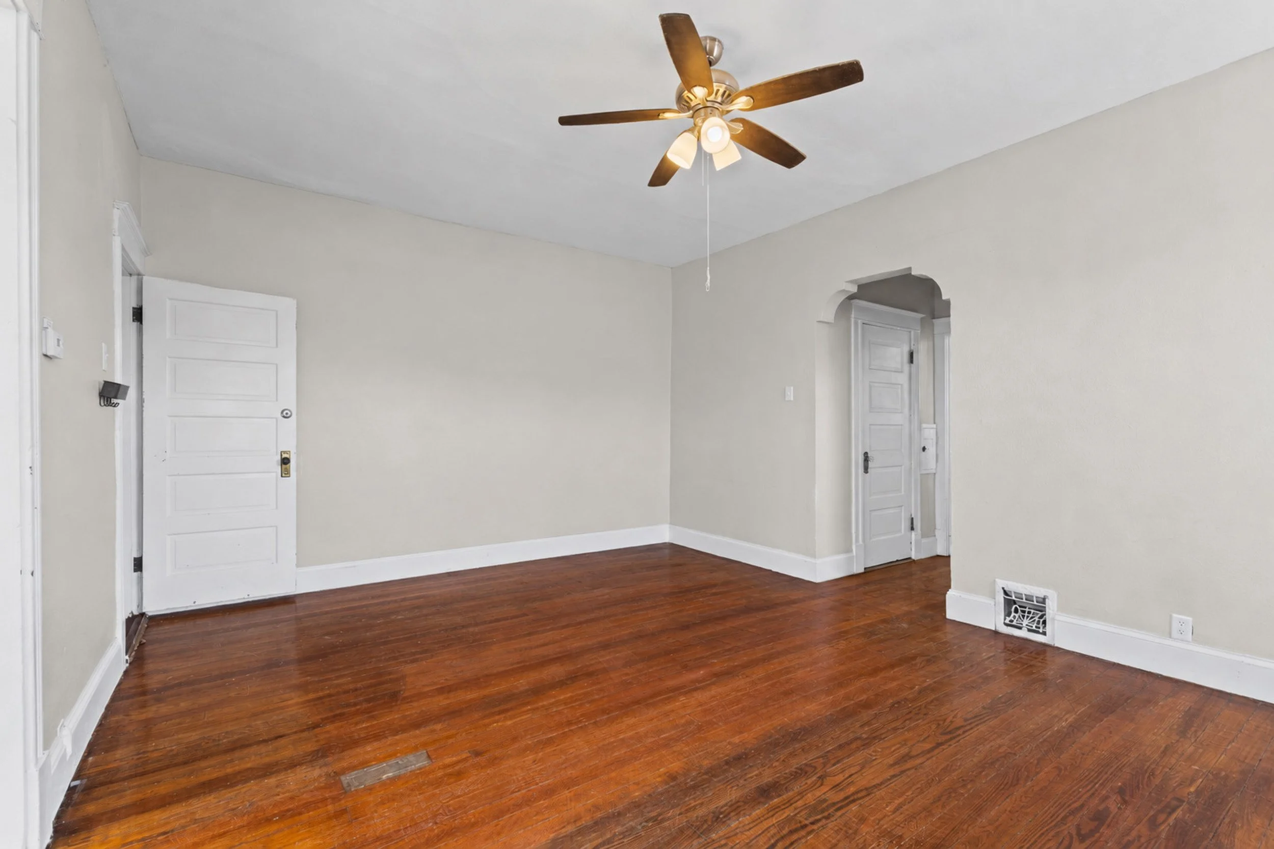 Empty room with hardwood floors, painted white walls, two white doors, a ceiling fan with wooden blades, and a ceiling light fixture.
