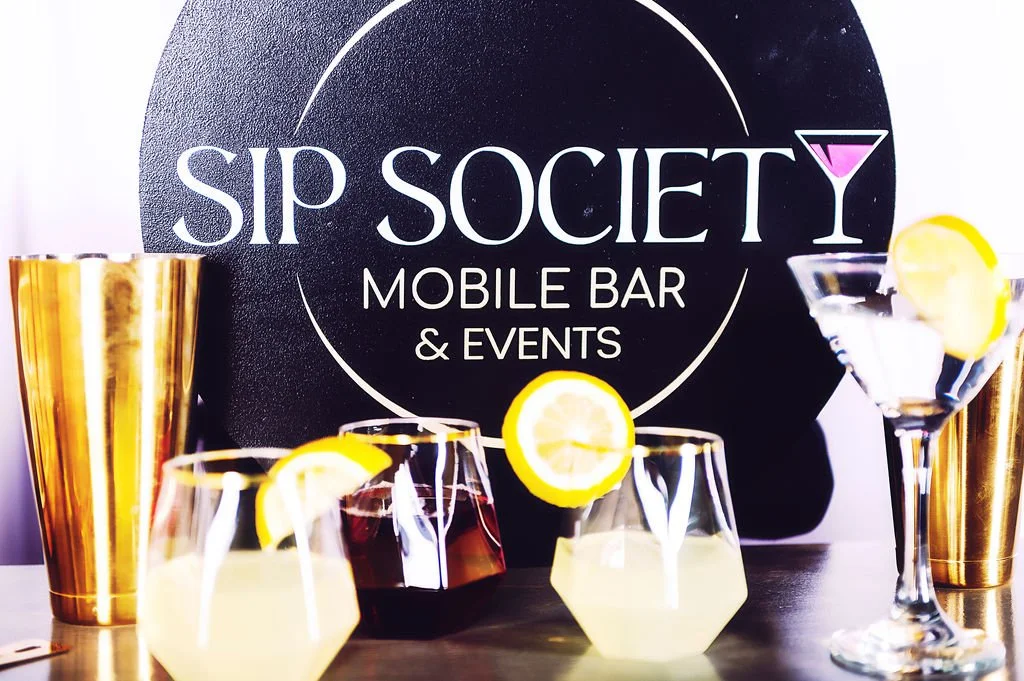A display of colorful cocktails with lemon garnishes in front of a sign that reads 'SIP SOCIETY MOBILE BAR & EVENTS' with gold cups on each side.