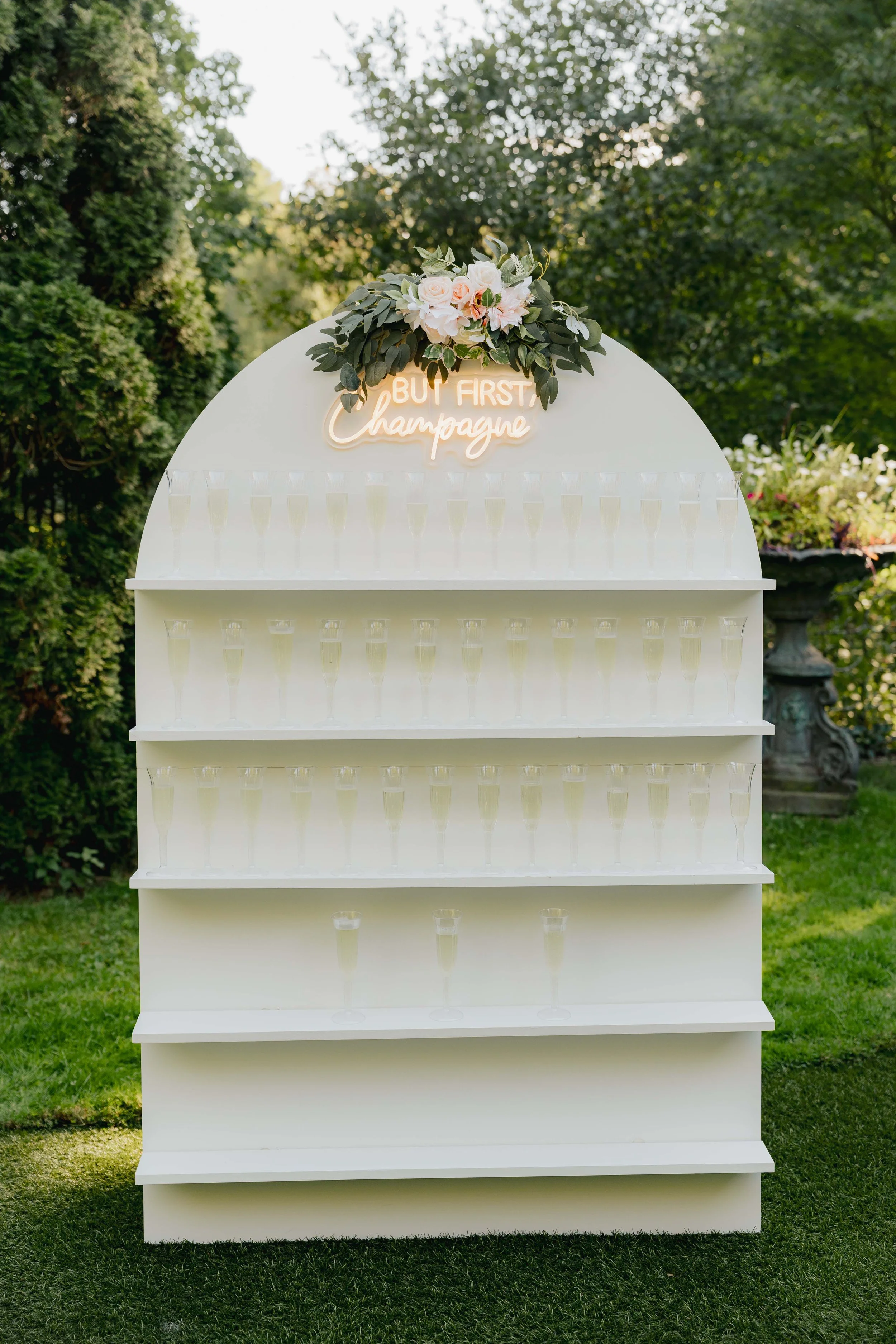 White shelf with multiple rows of champagne glasses, decorated with green foliage and pink flowers at the top, with a neon sign reading 'But First Champagne,' set outdoors on grass surrounded by greenery.