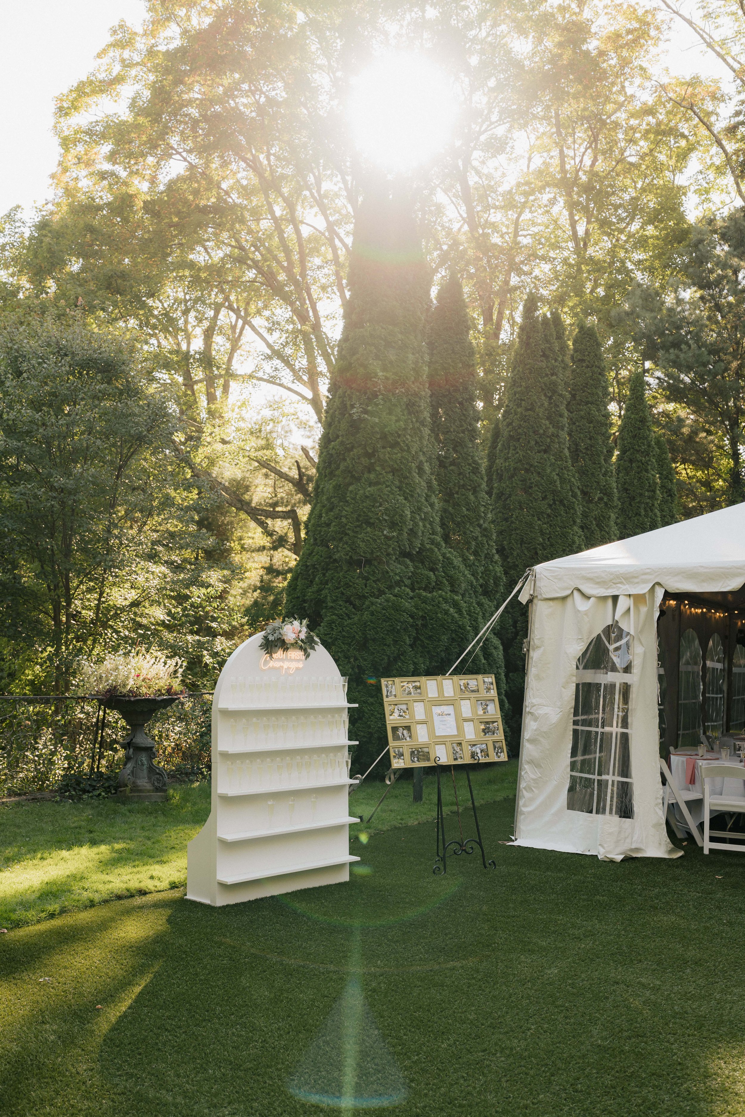 Outdoor event space with a white tent, picture frame display, and a white shelf decorated with flowers, surrounded by tall trees under sunlight.