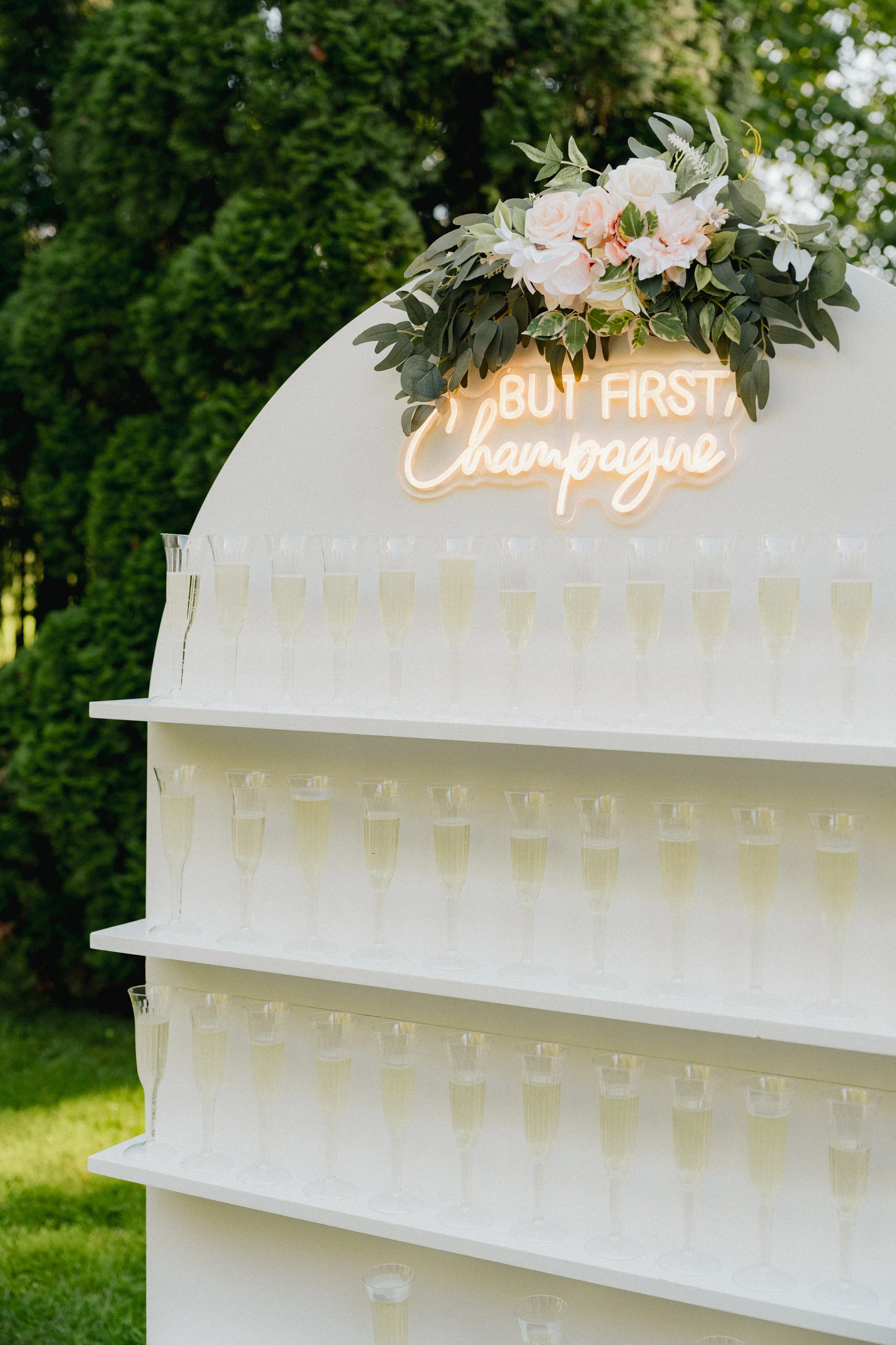 A white display stand with champagne glasses filled with champagne and a floral arrangement at the top. A neon sign reads 'But First, Champagne'.