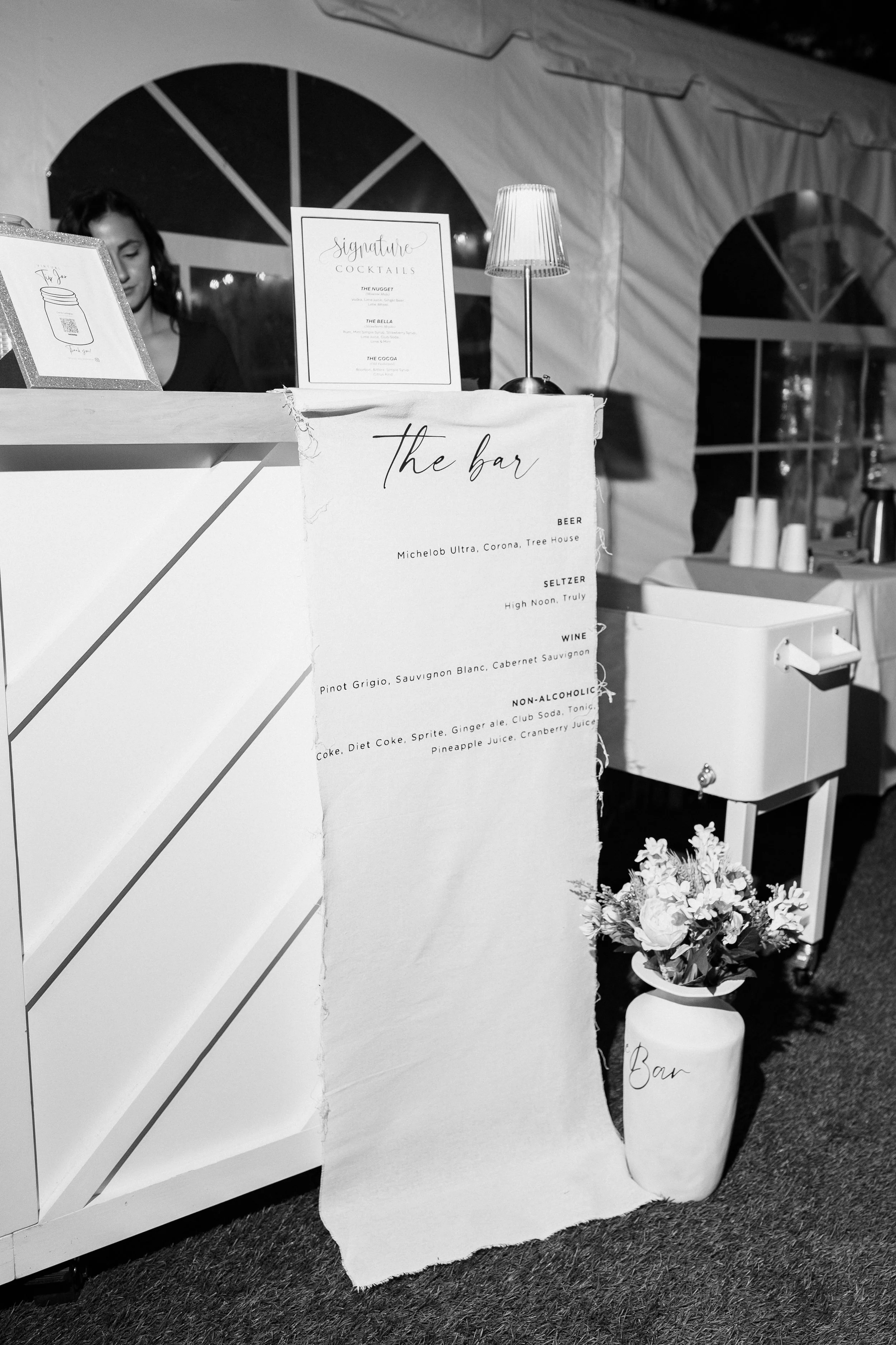 A black and white photograph of a bar setup at an event. There is a handwritten-style sign listing drink options including beer, seltzer, wine, and non-alcoholic beverages. A table or counter with a lamp, flowers in a vase labeled 'Bar', and a woman 