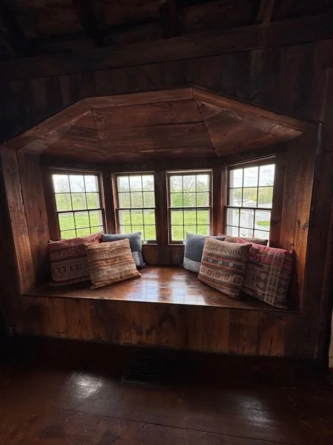 Wooden window seat with pillows, surrounded by multiple window panes with a green landscape view outside.