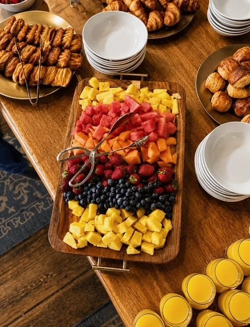 A wooden table with three platters of croissants, a fruit platter with watermelon, cantaloupe, pineapple, strawberries, and blueberries, and glasses of orange juice. There are stacks of white bowls around the table.