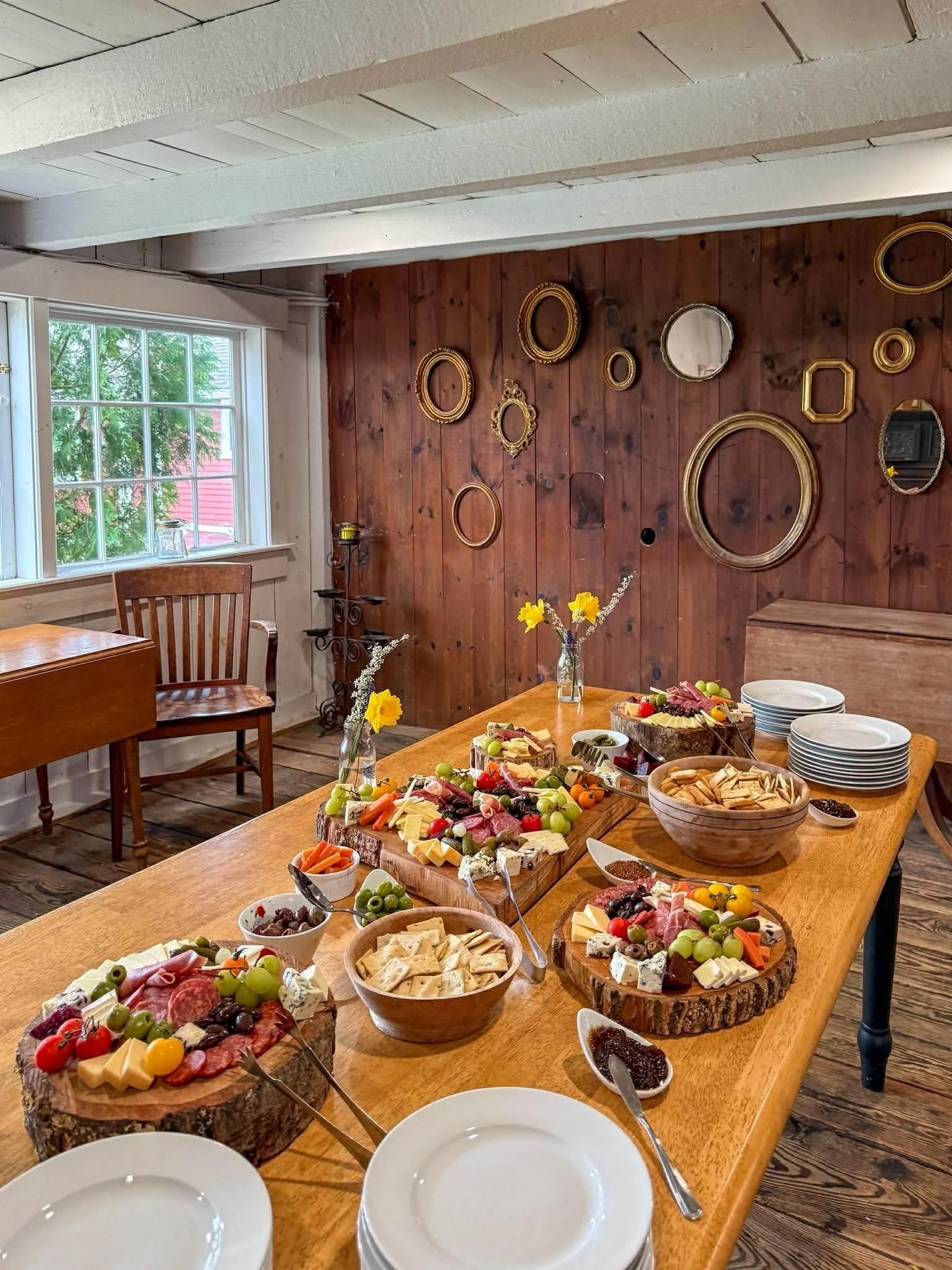 A dining table with various cheeses, meats, crackers, fruits, and spreads, set for a buffet. There are empty white plates in the foreground, and a wooden wall with multiple empty picture frames in the background, next to a window with a view of green