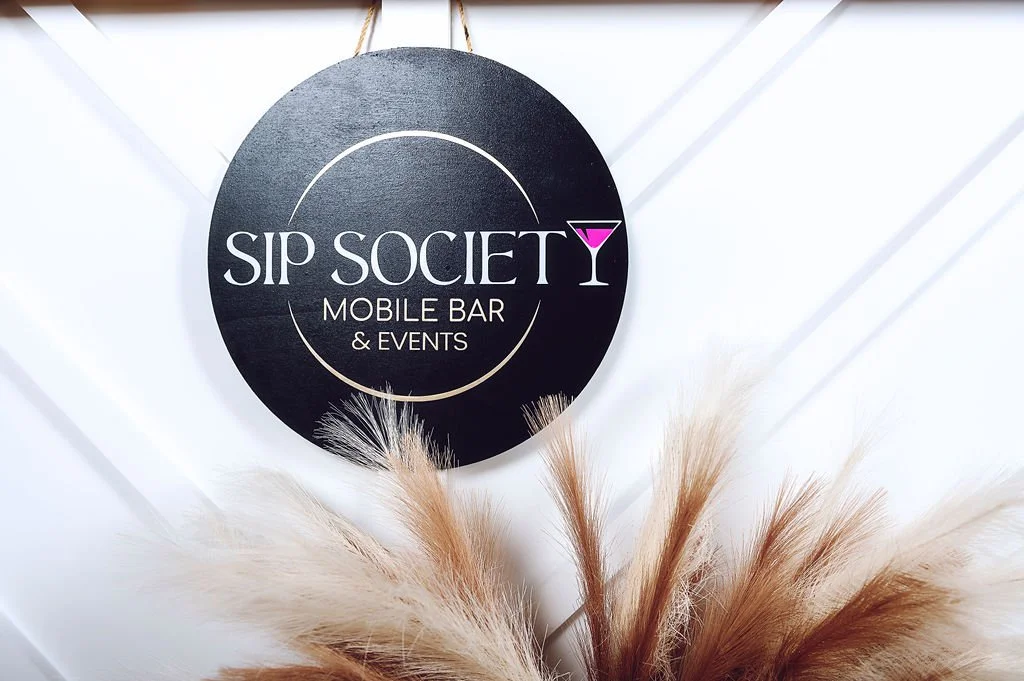 Black circular sign with the words 'SIP SOCIETY MOBILE BAR & EVENTS' and a pink martini glass graphic, hanging on a white wall with some decorative dried plants at the bottom.