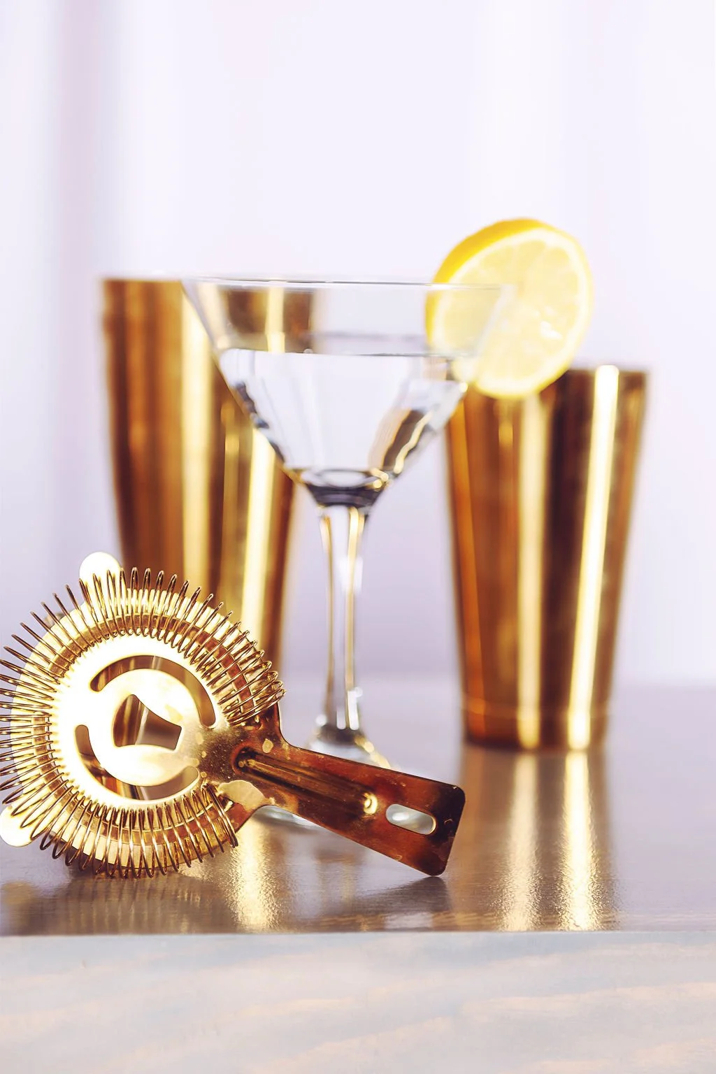 A cocktail glass with a lemon slice, gold cocktail shaker, and a gold cocktail strainer on a reflective surface.