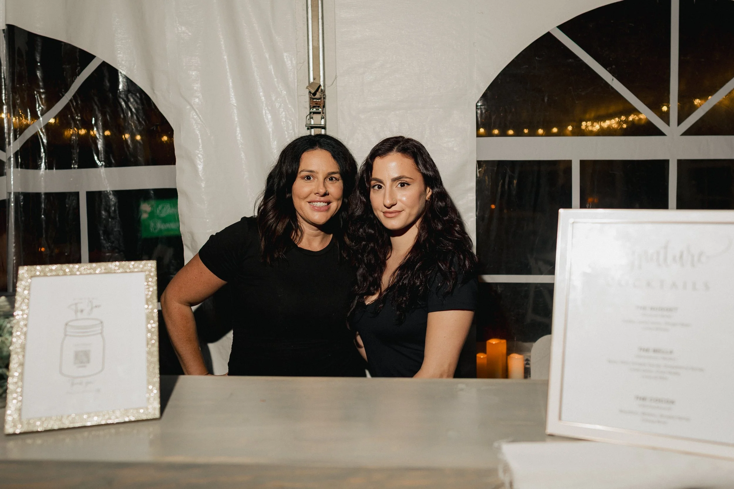 Two women standing behind a table at a night event, with menu boards and candles in the background.