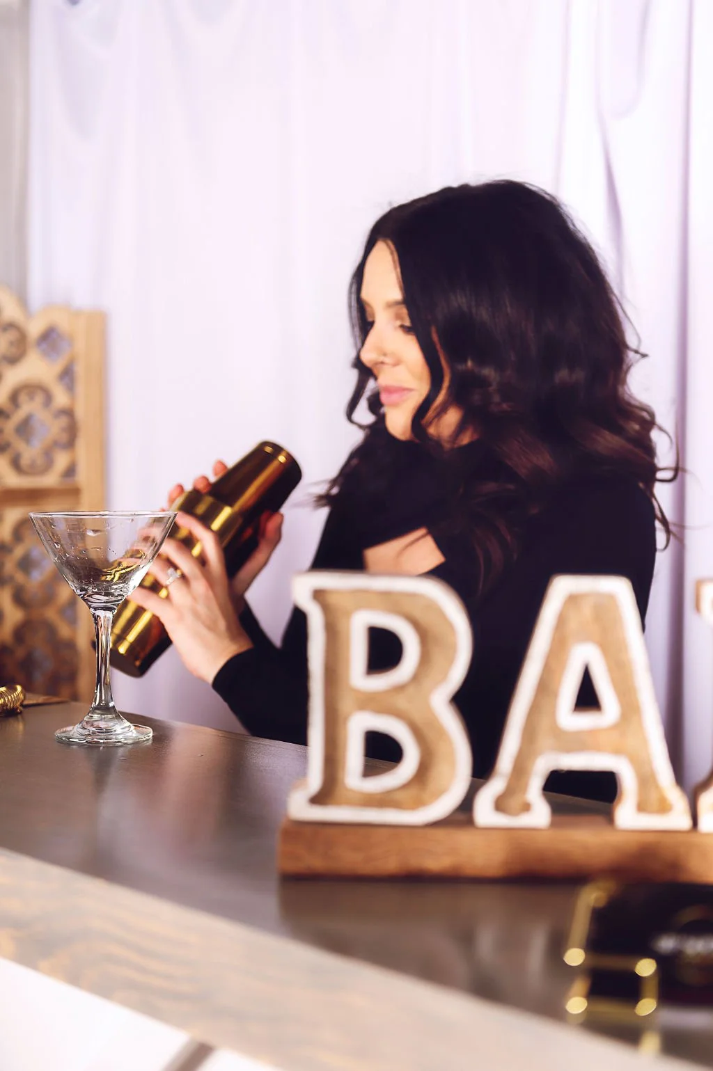 A woman with dark hair, dressed in black, stands behind a bar counter with a cocktail glass and a shaker. Decor in the background and a sign with the word 'BAR' in large letters are visible.