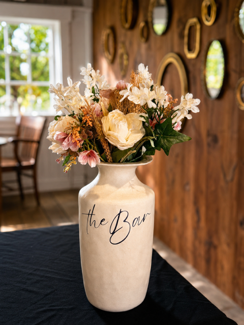 Vase with a floral arrangement of white, pink, and peach flowers, labeled "the Bar" in black script, on a black table in a cozy room with a wooden wall and circular mirrors.