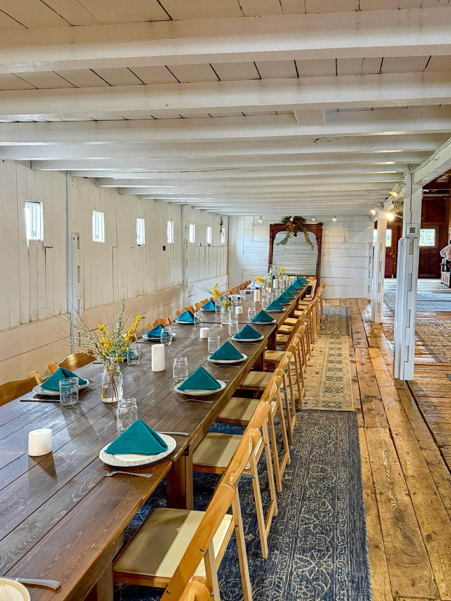 Long wooden dining table set with teal napkins, glassware, and white candles. Floral arrangements decorate the center of the table. The room has white paneled walls, a wooden ceiling, and a decorative rug underneath the table. In the background, a wo