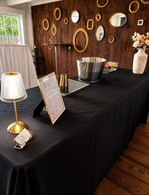 A black-covered table with a white lamp, a gold drinking cup, a framed sign, a large ice bucket, and a white vase with pink flowers in a decorated room with wooden walls and a wall of circular mirrors.
