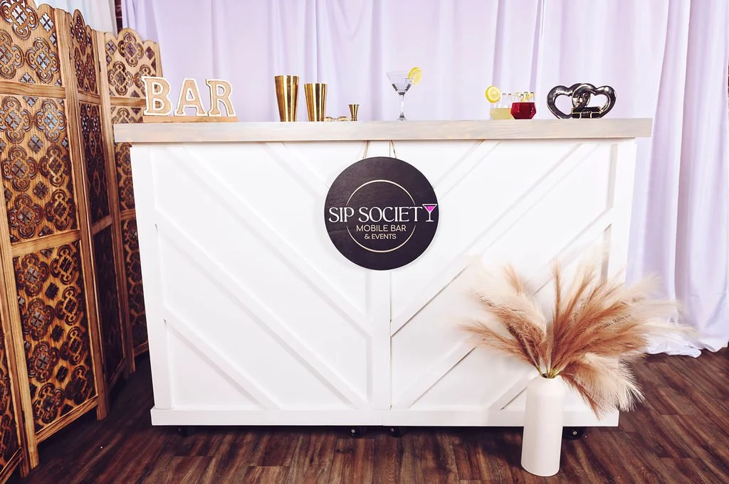 A white bar counter with a black circular sign that says "Sip Society Mobile Bar & Events". On top of the counter are gold cups, cocktail glasses with drinks, and a black heart-shaped decorative piece. To the left, a wooden decorative folding screen. To the right, a white vase with pampas grass. The background has white curtains.