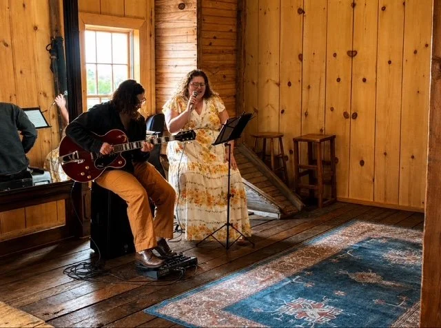 Two women performing music in a wooden interior, one playing guitar and the other singing into a microphone, with a stereo and a window in the background.