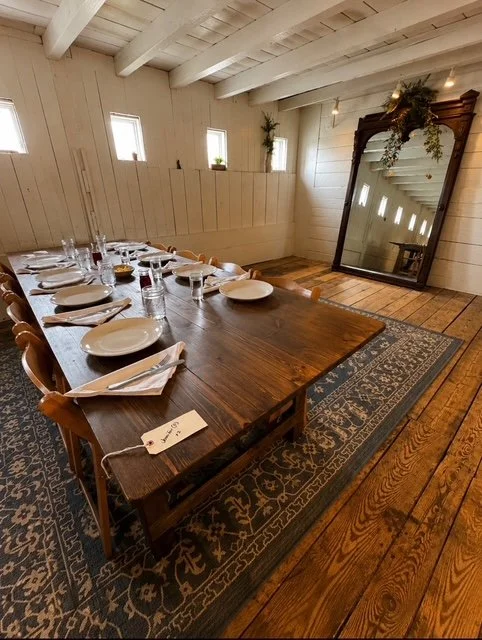 A dining room with a large wooden table set with plates, glasses, and napkins, a patterned rug, and a large mirror leaning against the wall.