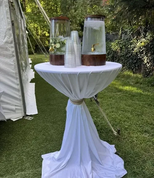 Two large beverage dispensers with lemon slices and mint leaves, placed on a round table with a white tablecloth, outdoor setting with grass and trees.