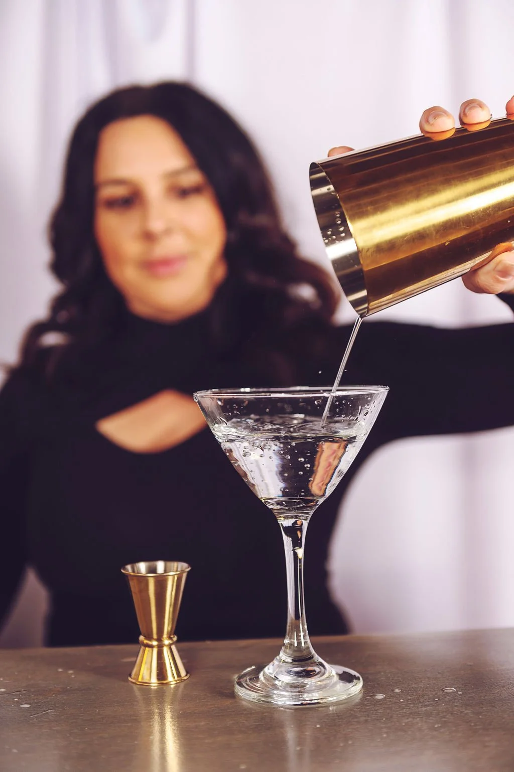 A woman with dark hair in a black top is pouring a clear liquid from a gold cocktail shaker into a martini glass, with a gold jigger on the table.