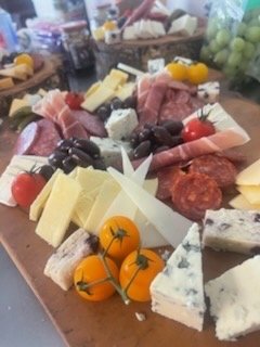A wooden cheese board with various cheeses, cured meats, grape tomatoes, and small yellow tomatoes, set in a grocery or deli setting.