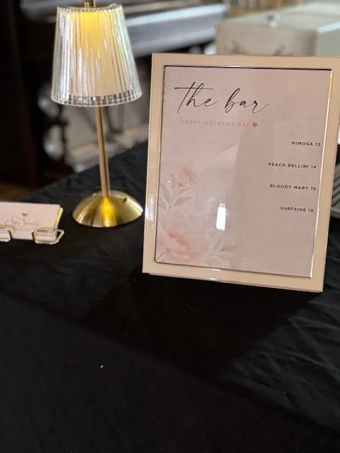 A framed sign on a table with a black tablecloth, titled 'the bar' with a Mother's Day message, listing four drink options. A small table lamp with a gold base and silver shade is nearby.