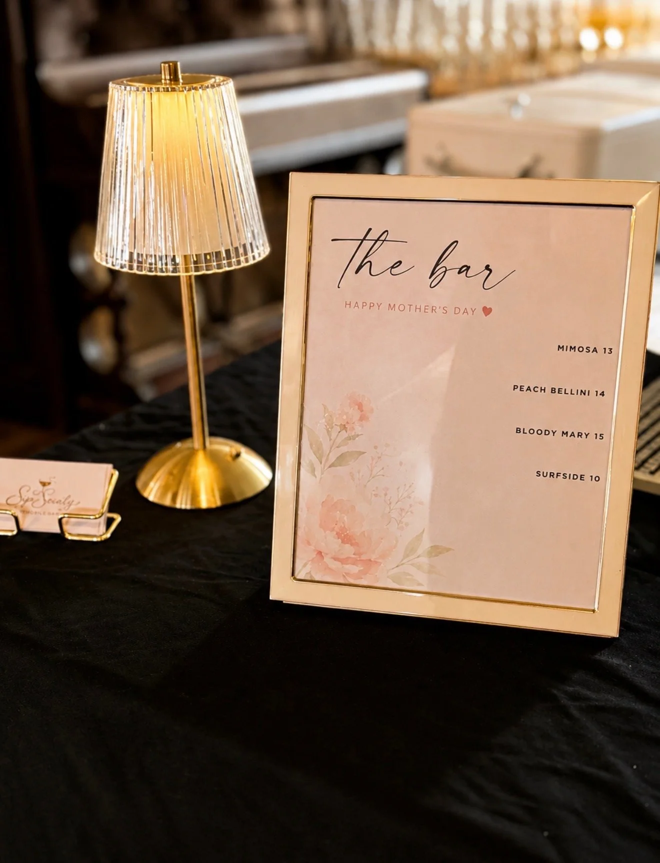 A golden table lamp with a ribbed, transparent shade sits next to a framed sign that reads 'the bar' with a Mother's Day greeting and a list of drinks and prices, on a black tablecloth.